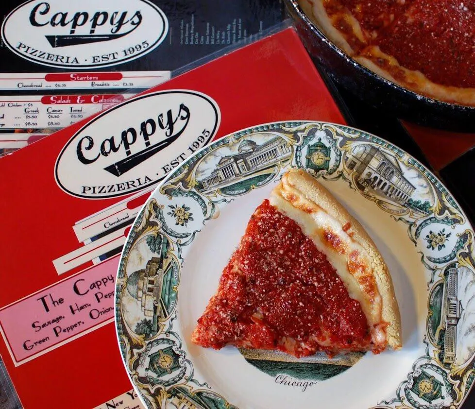 A slice of Cappy's Chicago style deep dish pizza on a Chicago souvenir plate, with the full pan behind it